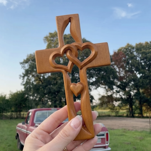 Hand holding a wooden cross with heart designs against a scenic background with trees and a truck.